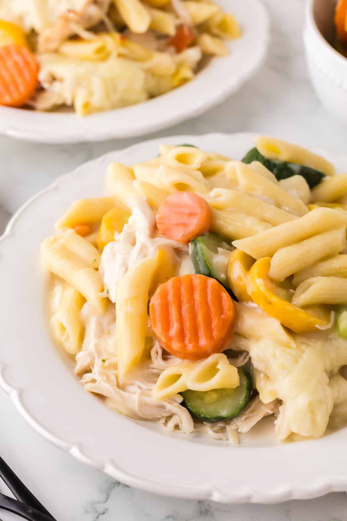 A white plate filled with chicken vegetable pasta—penne, sliced carrots, zucchini, and shredded chicken in a creamy sauce. Another similar plate is blurred in the background on a white marble surface.