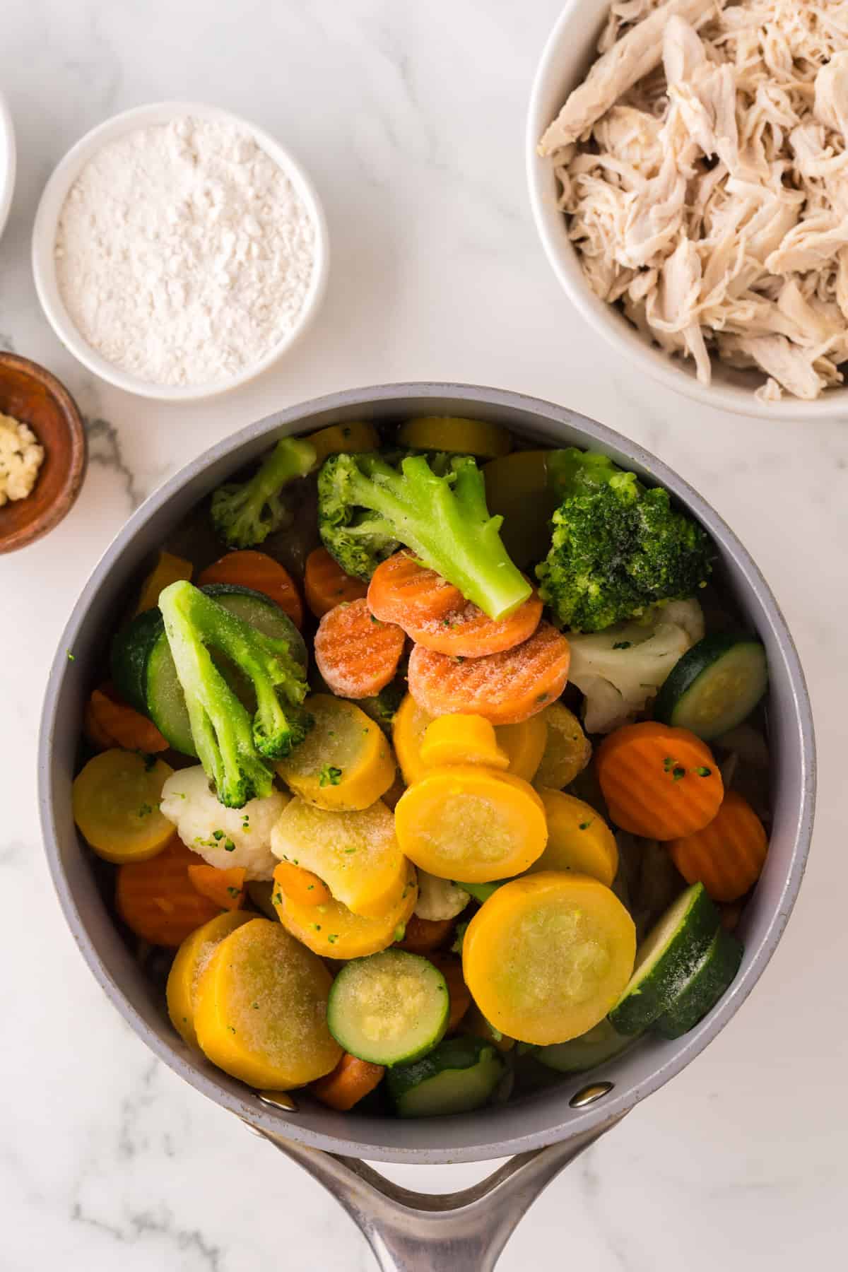 A saucepan filled with sliced carrots, zucchini, broccoli, and yellow squash sits on a marble surface beside bowls of shredded chicken, flour, minced garlic, and chicken vegetable pasta ingredients.
