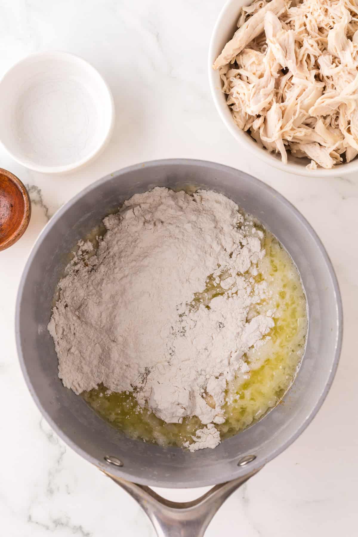 A saucepan on a marble countertop contains melted butter and flour. Nearby, there is a bowl of shredded chicken, a small white bowl, a wooden container, and hints of chicken vegetable pasta ready to be prepared.