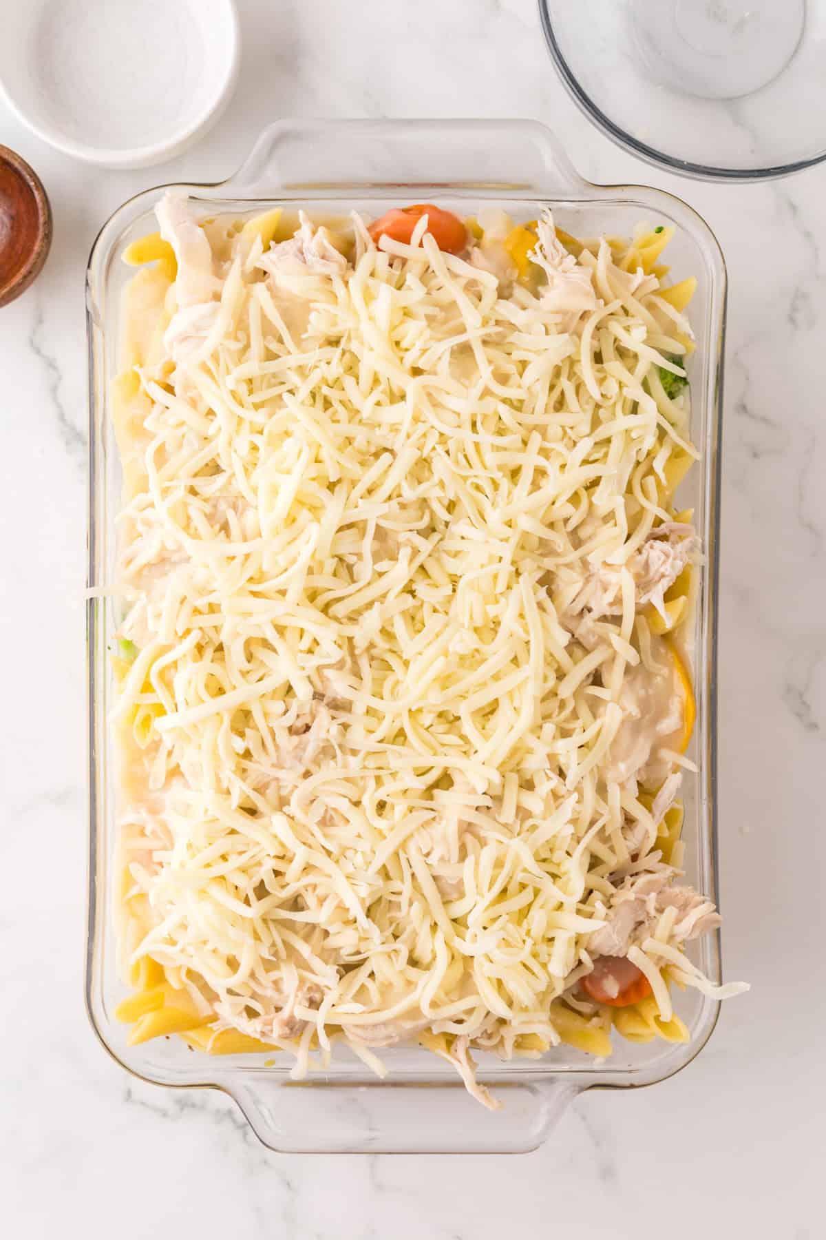 A glass baking dish filled with an unbaked chicken vegetable pasta casserole, topped with shredded cheese and featuring visible pasta and vegetables beneath the golden cheese layer, set on a white marble countertop.