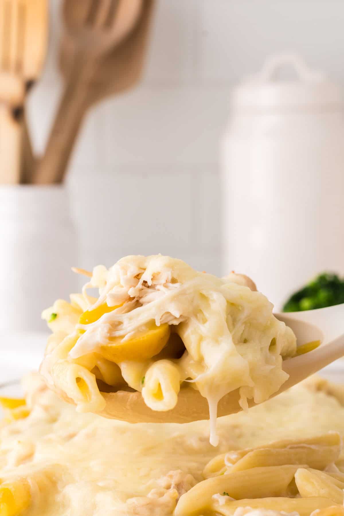 A close-up of a spoon lifting a serving of cheesy chicken vegetable pasta with penne, shredded chicken, veggies, and melted cheese from a casserole dish. Wooden utensils and kitchen containers are blurred in the background.