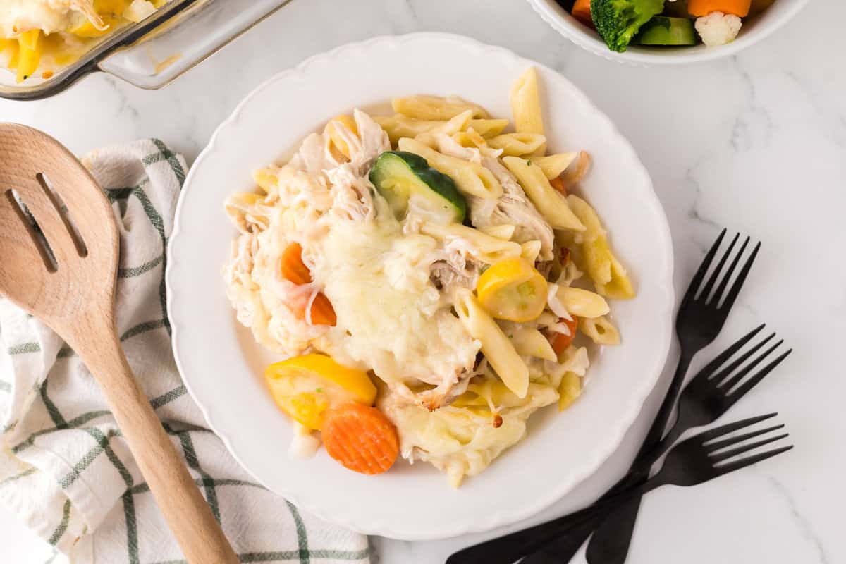 A white plate with cheesy chicken vegetable pasta—baked penne, shredded chicken, and mixed veggies like carrots, zucchini, and squash—sits beside a napkin, wooden utensils, and three black forks on a white table.
