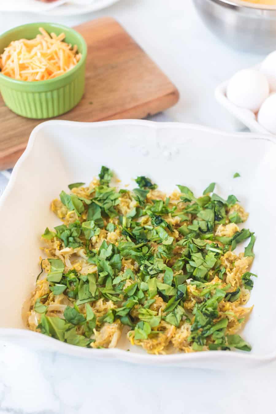 A white baking dish filled with a mixture of sautéed vegetables and fresh chopped spinach, perfect for an egg bake. Nearby, there are eggs, shredded cheese in a green bowl, and a wooden cutting board on a white countertop.
