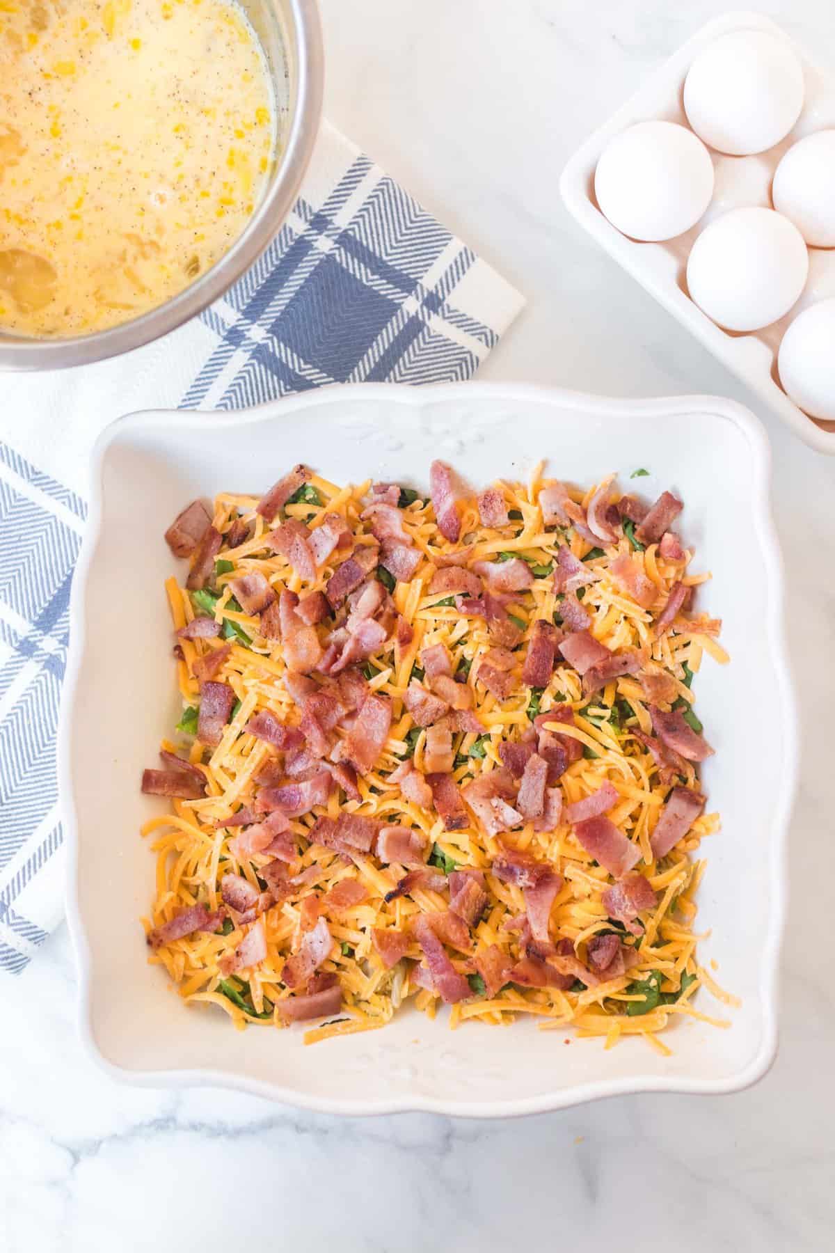 A white baking dish with chopped bacon, shredded cheese, and greens sits on a marble surface, ready for an egg bake. Nearby are a bowl of whisked eggs, a blue-and-white towel, and a carton of eggs.
