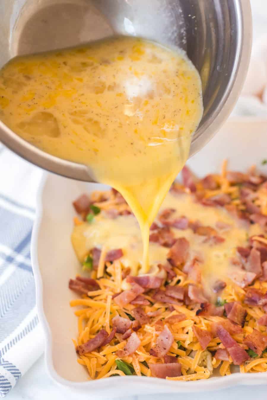 A bowl of beaten eggs is being poured over a casserole dish filled with shredded cheese, chopped bacon, and vegetables, creating a savory egg bake ready for the oven.