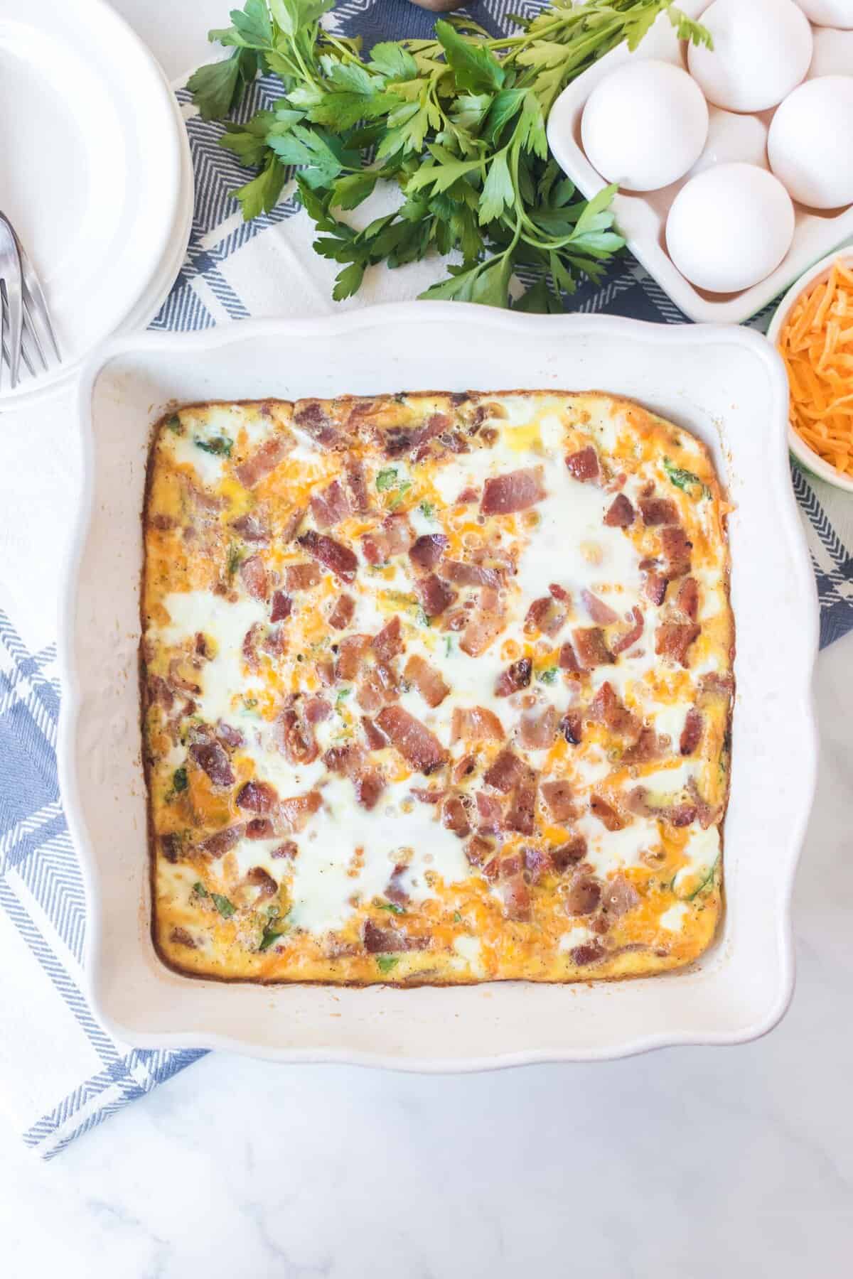 A savory egg bake casserole with bacon, cheese, and herbs in a square white dish, surrounded by fresh parsley, whole eggs, shredded cheese, and a stack of plates on a blue and white cloth.