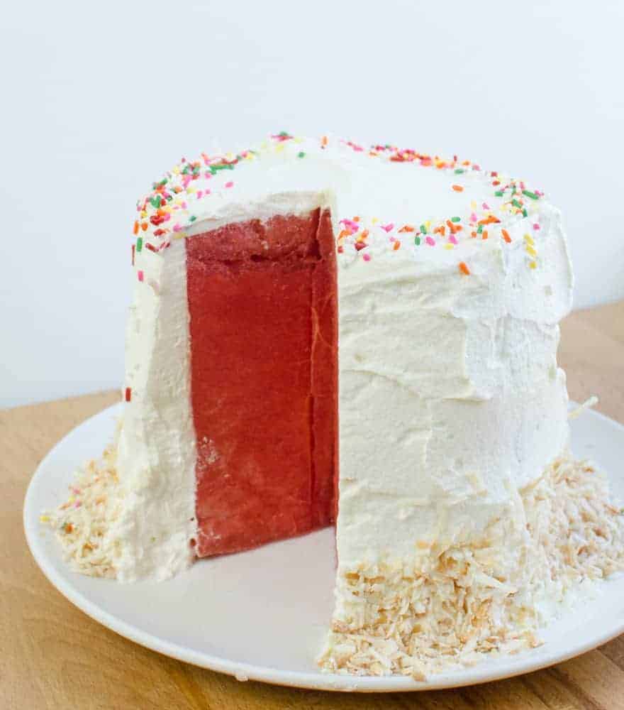 Watermelon Cake with Whipped Cream frosting on a white plate decorated with sprinkles and toasted coconut.