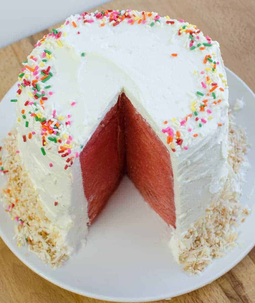 Top view of a Watermelon Cake with Whipped Cream frosting on a white plate decorated with sprinkles and toasted coconut.