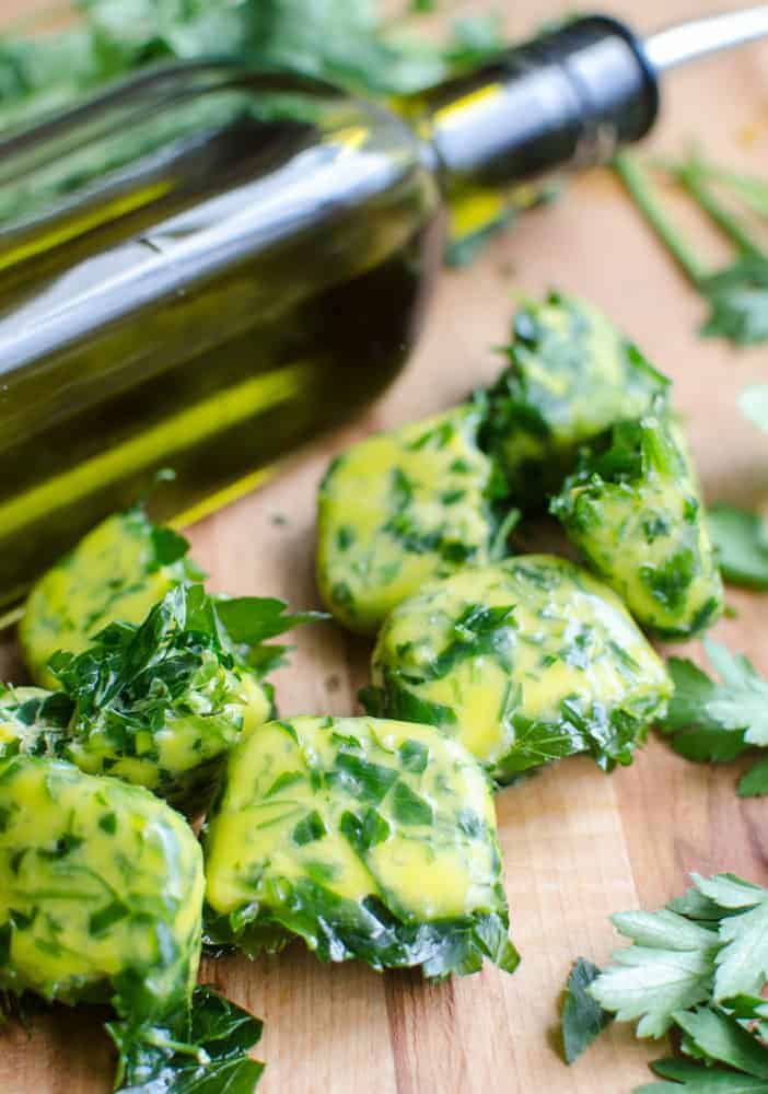 Frozen herb bombs on a wooden cutting board with a bottle of olive oil in the background and some fresh herbs next to them.