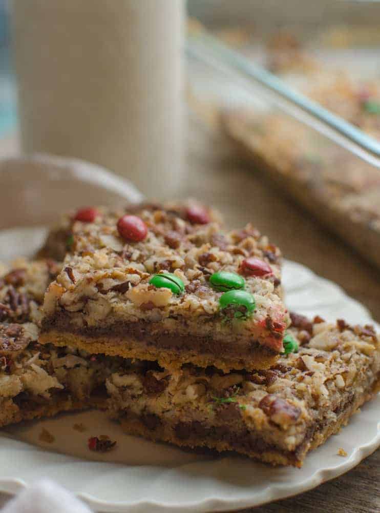 Three square Christmas Magic Cookie Bars stacked in a pile on a white plate with the full pan of bars in the background.