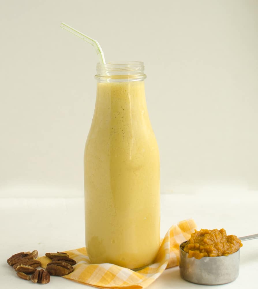 Pumpkin Protein Smoothie in a glass bottle with a straw sitting on a yellow and white gingham napkins with some pecans and a measuring cup of pumpkin puree.