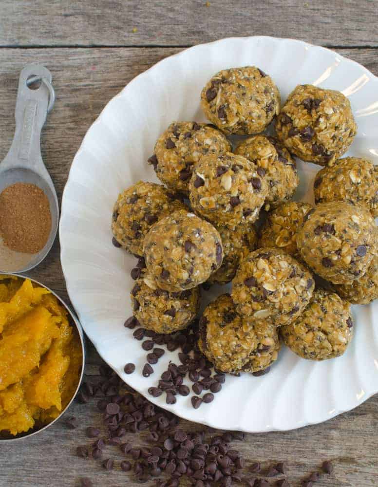 A white plate holds a pile of round, textured oatmeal energy bites with visible oats and chocolate chips. Next to it, a measuring spoon contains cinnamon, and a bowl holds mashed pumpkin. More chocolate chips are scattered on the wooden surface.