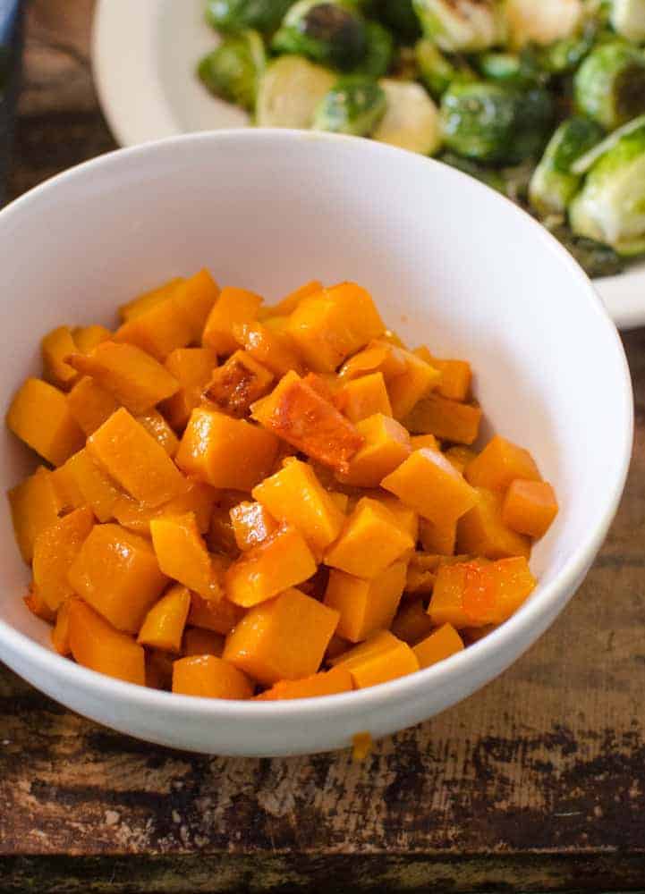 A white bowl filled with small, evenly cut cubes of cooked butternut squash sits on a wooden surface, making it a delicious addition to thanksgiving dinner side dishes, with a plate of Brussels sprouts visible in the background.