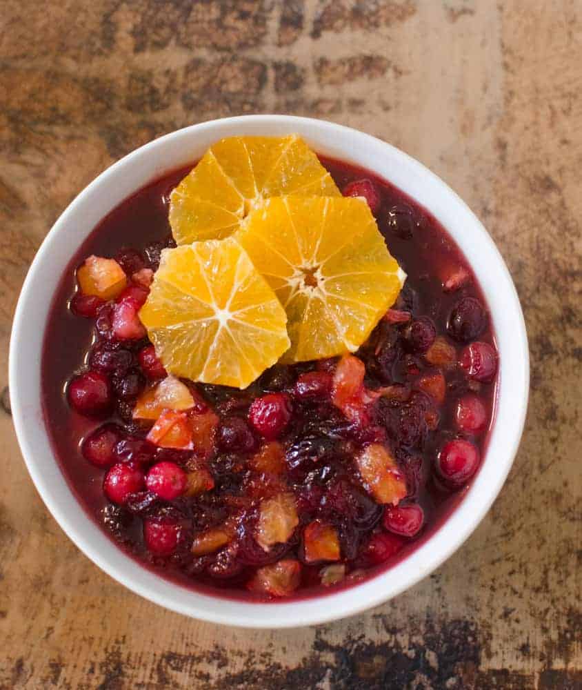 A white bowl filled with cranberry sauce, containing whole cranberries and orange pieces, topped with three round slices of orange—an inviting addition to your thanksgiving dinner side dishes on a rustic wooden surface.