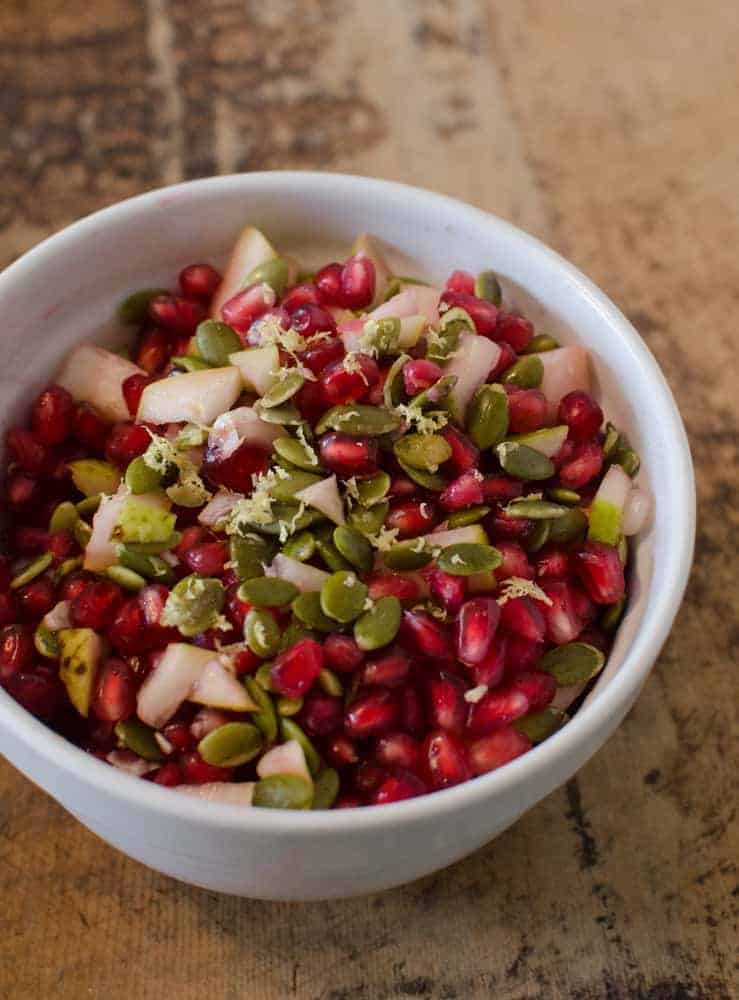 A white bowl filled with a colorful salad of pomegranate seeds, green pumpkin seeds, diced pear, and a sprinkle of lemon zest sits on a rustic brown surface—perfect for your Thanksgiving dinner side dishes.