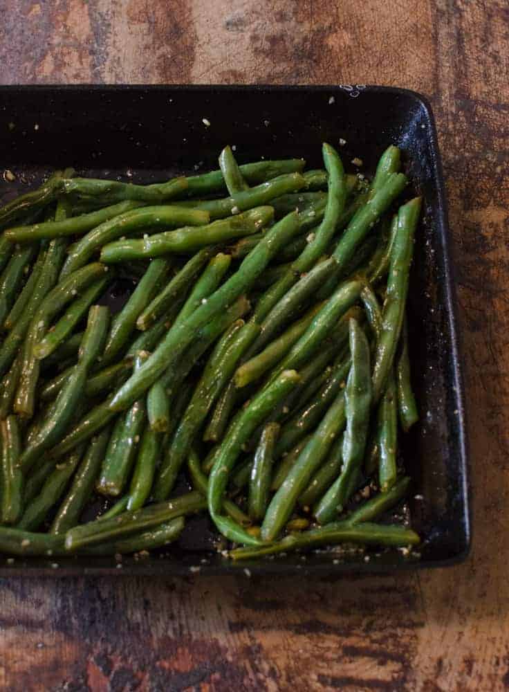 A black rectangular dish filled with cooked green beans, lightly coated in seasoning, sits on a rustic brown tabletop—an inviting choice among classic Thanksgiving dinner side dishes.