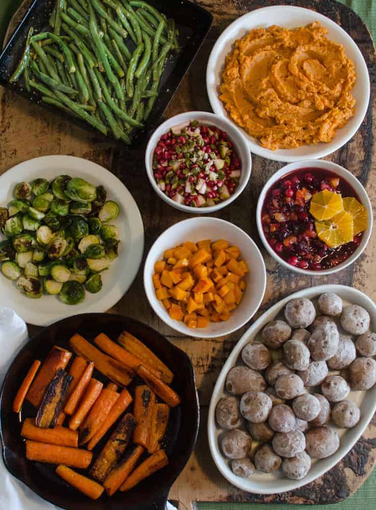 A vibrant display of Thanksgiving dinner side dishes, including green beans, mashed sweet potatoes, Brussels sprouts, roasted carrots, pomegranate salsa, cranberry-orange sauce, diced squash, and roasted baby potatoes.
