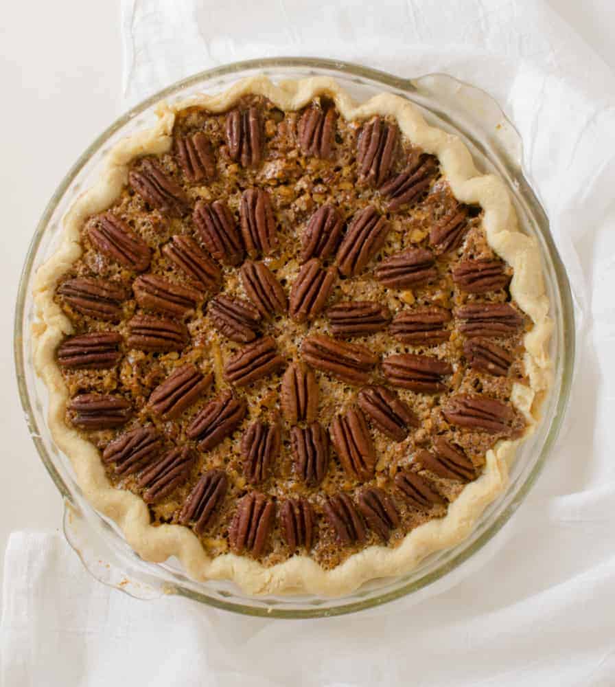 A whole pecan pie in a glass dish, featuring a golden, flaky crust and a decorative pattern of whole pecans on top of the filling, placed on a white cloth—perfect inspiration for your collection of 8 pie recipes.