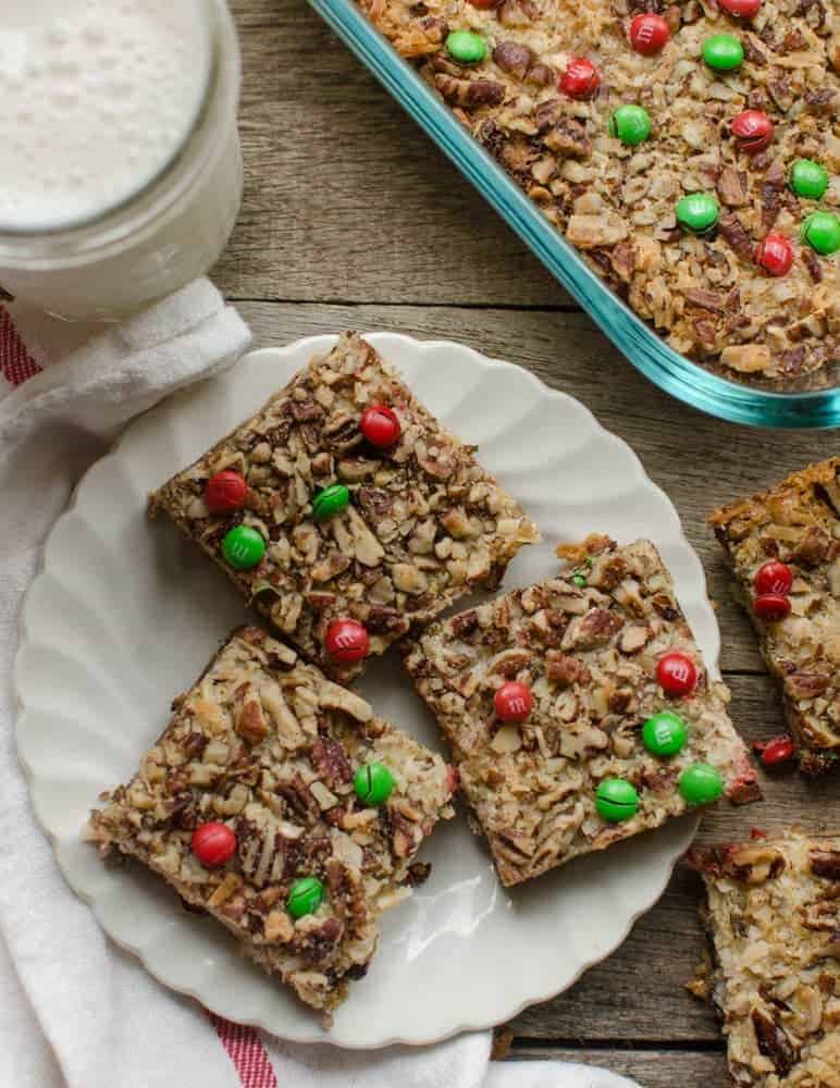 Three Christmas Magic Cookie Bar squares on a white plate next to a glass of milk and a full pan of bars.