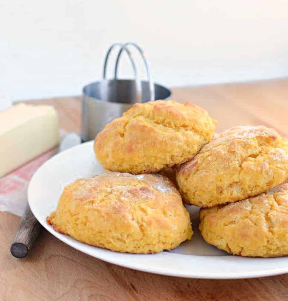 Four Sweet Potato Biscuits on a white plate next to a sticker of butter and a knife on a butcher block counter.