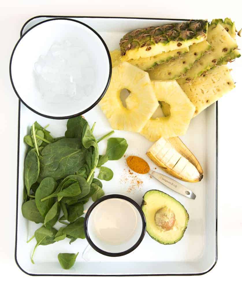The ingredients for a workout recovery smoothie on a white enamelware pan with a black rim, including a bowl of ice, a pile of fresh baby spinach, a couple of pineapple rings, half of a banana, half an avocado, a measuring spoon with ground tumeric, and a white cup with almond milk.