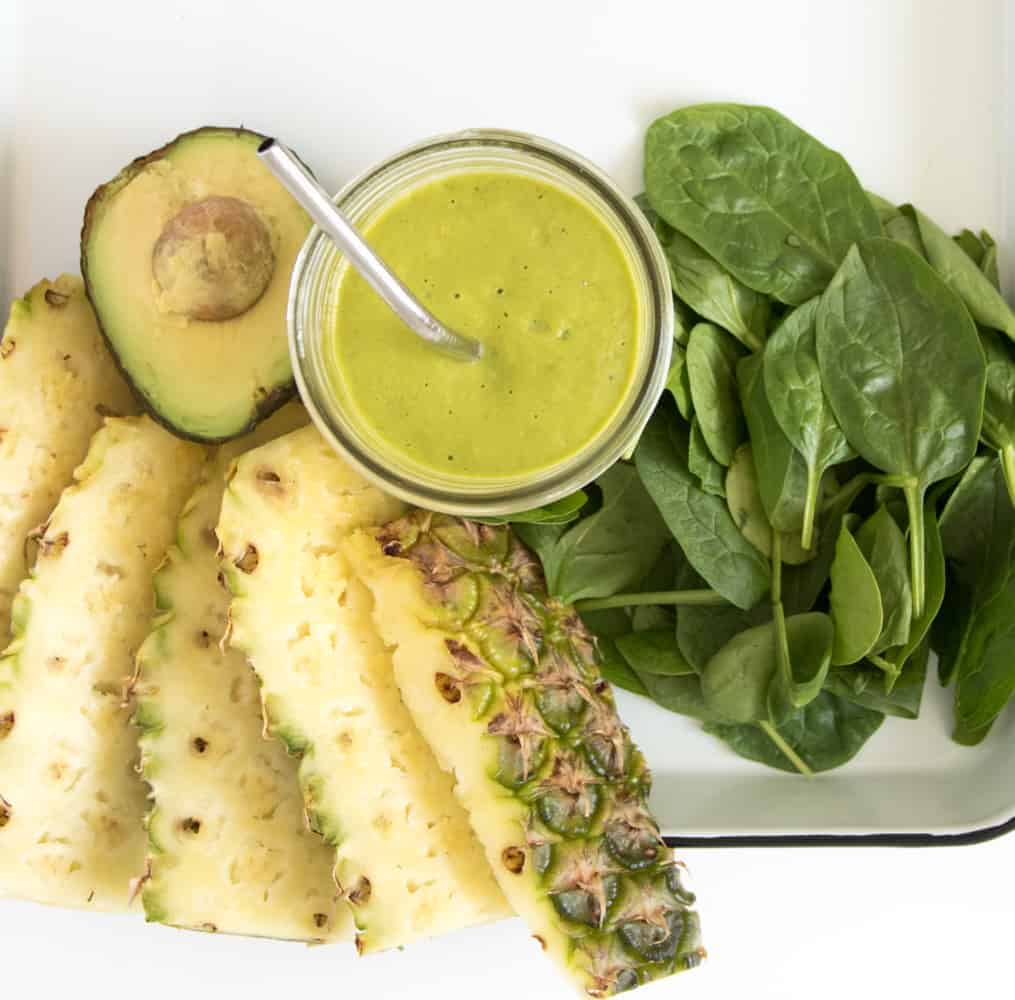 Top view of a workout recovery smoothie in a mason jar with a straw next to half an avocado, some pineapple slices, and some fresh spinach on a white enamelware pan with a black rim.
