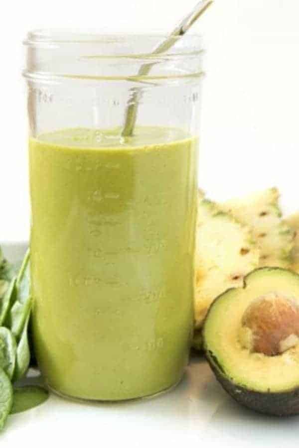 A workout recovery smoothie in a mason jar with a straw next to half an avocado, some pineapple slices, and some fresh spinach.