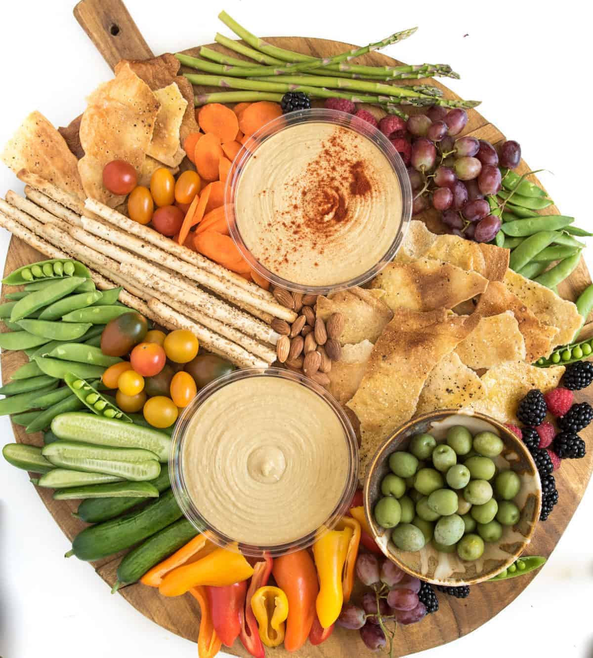 A round wooden hummus platter features two bowls of hummus—one topped with paprika—surrounded by breadsticks, crackers, olives, grape tomatoes, cucumber slices, snap peas, grapes, berries, nuts, mini bell peppers, carrots, and asparagus.