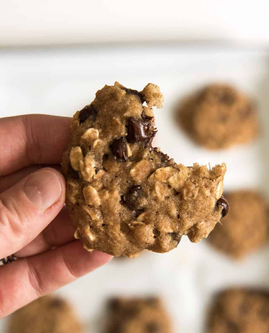 A hand holding an applesauce oatmeal cookie with a bite taken out of it; more cookies are blurred in the background on a baking sheet.