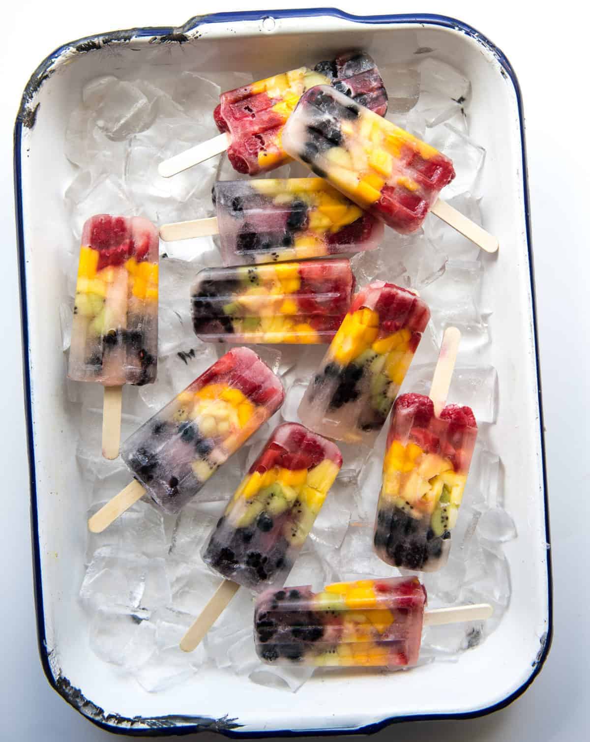 10 healthy rainbow popsicles on a bed of ice in a white rimmed enamelware pan with a black rim.