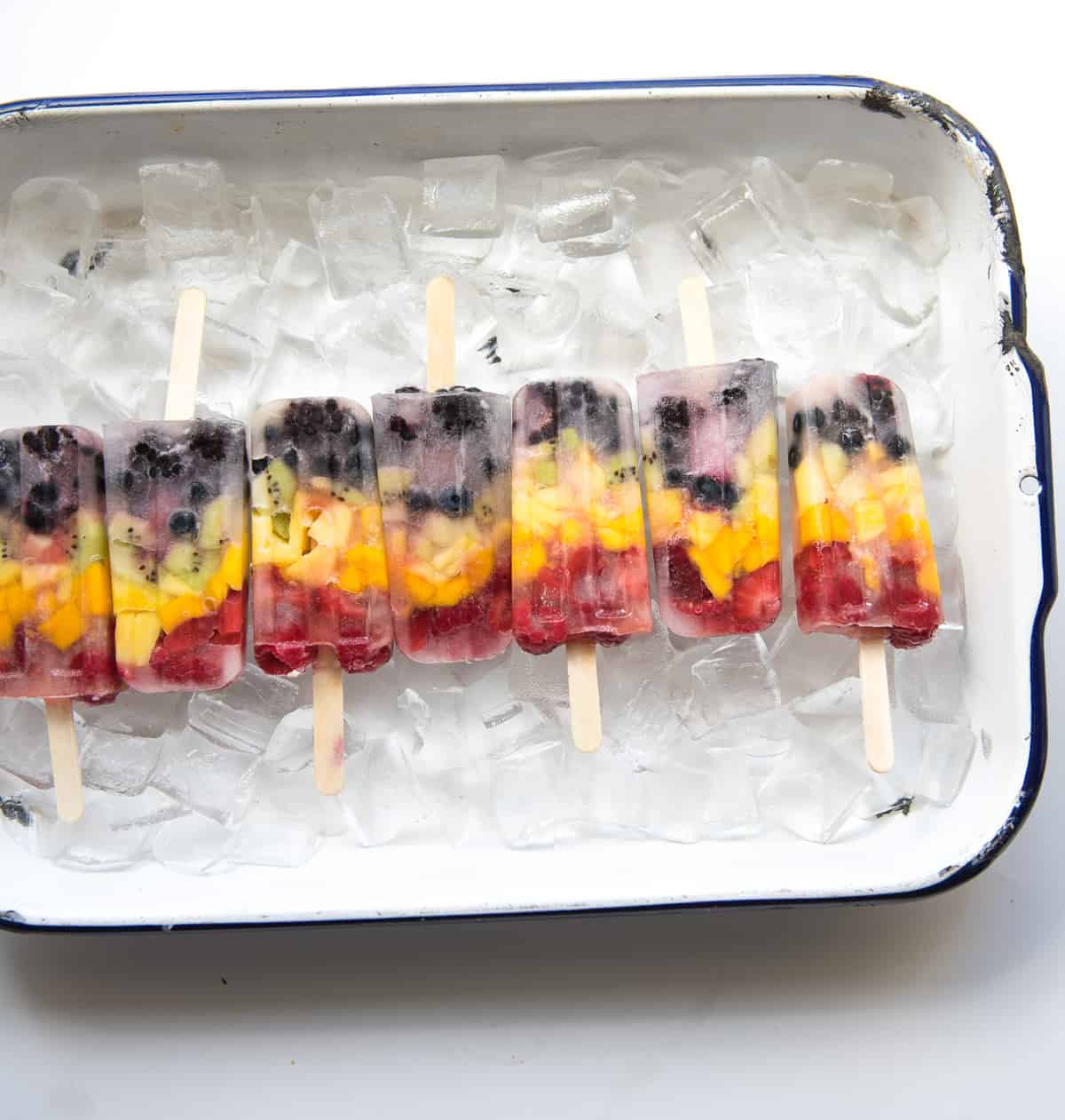 7 healthy rainbow popsicles lined up with sticks pointing opposite directions on a bed of ice in a white enamelware pan with a black rim.