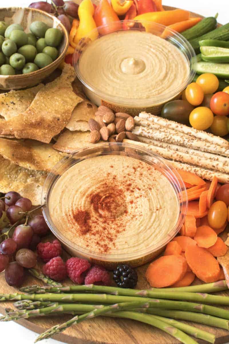 A vibrant hummus platter featuring two containers of hummus, assorted crackers, pita chips, olives, grape tomatoes, sliced carrots, bell peppers, cucumber, nuts, grapes, raspberries, a blackberry, and asparagus spears.