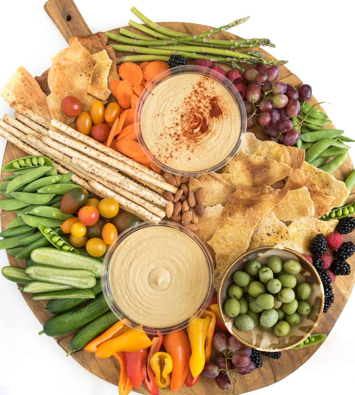 A round wooden hummus platter topped with two bowls of hummus, a bowl of green olives, crackers, breadsticks, cherry tomatoes, snap peas, cucumber slices, mini bell peppers, asparagus, grapes, almonds, blackberries, and carrots.