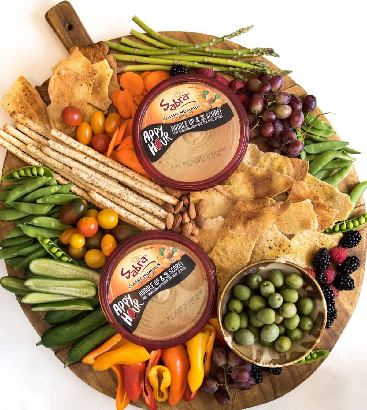 A round wooden board filled with colorful vegetables, breadsticks, crackers, olives, nuts, grapes, and two containers of Sabra hummus in the center creates a vibrant hummus platter arranged neatly for serving.