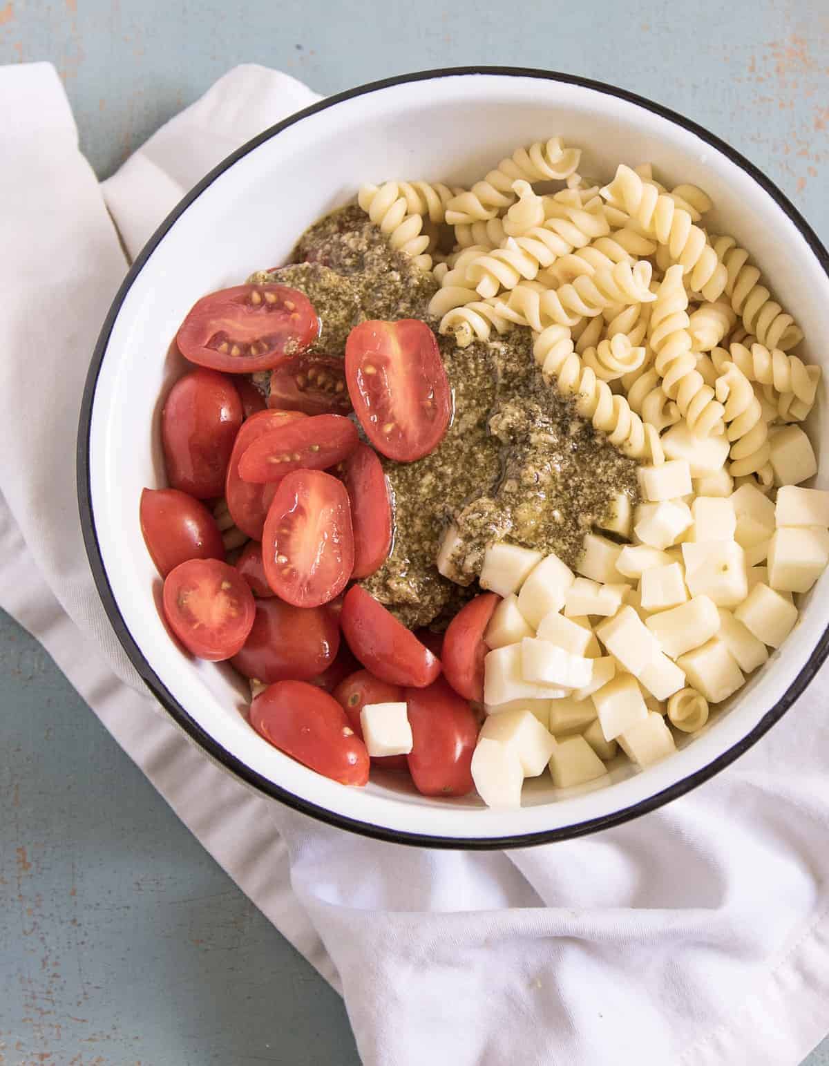 Ingredients for the Quick & Easy Pesto Pasta Salad in a white enamelware bowl with a black rim resting on a white napkin.