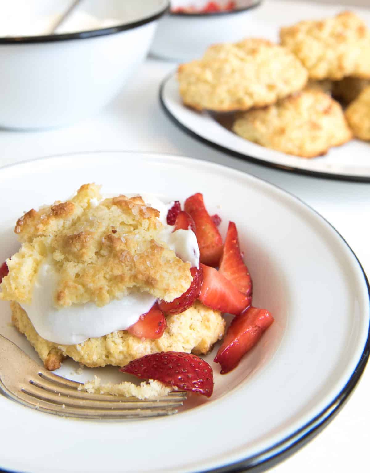 A white plate with strawberry shortcake, featuring sliced strawberries and whipped cream between two halves of a biscuit, with a fork beside it. A stack of biscuits is visible on a plate in the background.