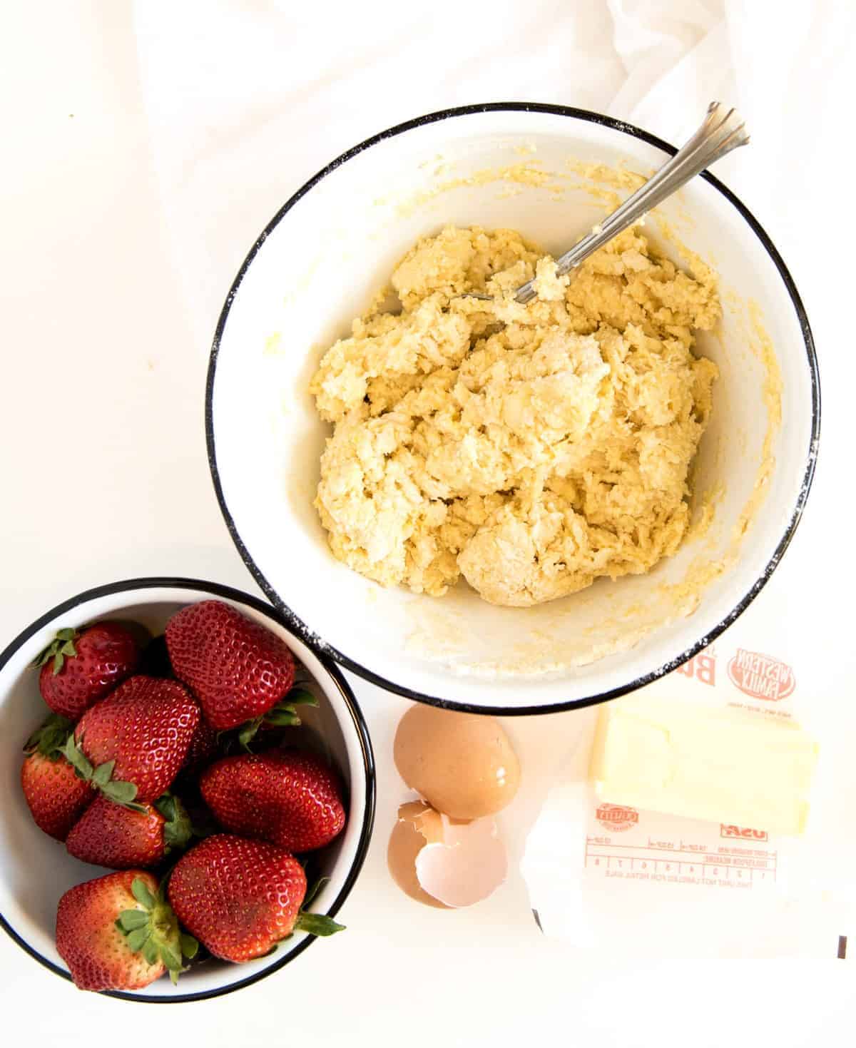 A bowl of dough with a spoon sits next to a bowl of fresh strawberries, ready to create the best strawberry shortcake. Nearby are two eggs, one cracked, and a stick of butter wrapped in paper, all resting on a pristine white surface.