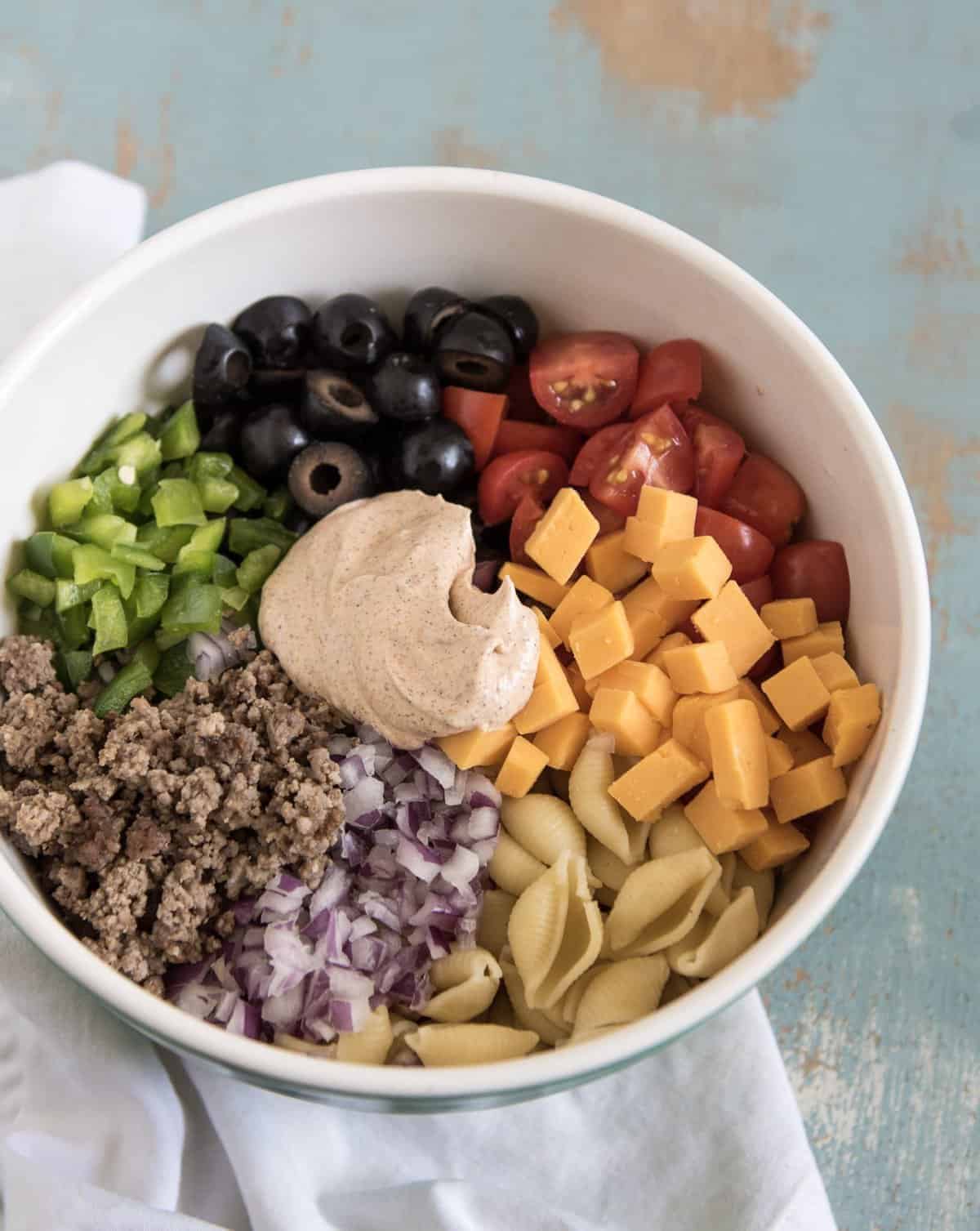 Ingredients for Creamy Taco Pasta Salad in a white bowl, including ground beef, bell peppers, black olives, grape tomatoes, cheddar cheese, shell pasta, red onions, and a dollop of the dressing.