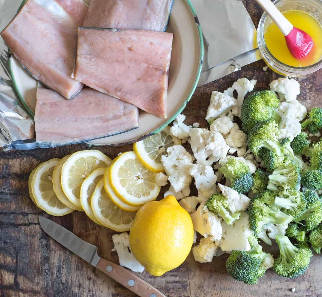 Top view of raw salmon on a plate next to a cutting board of lemon, cauliflower, and broccoli.