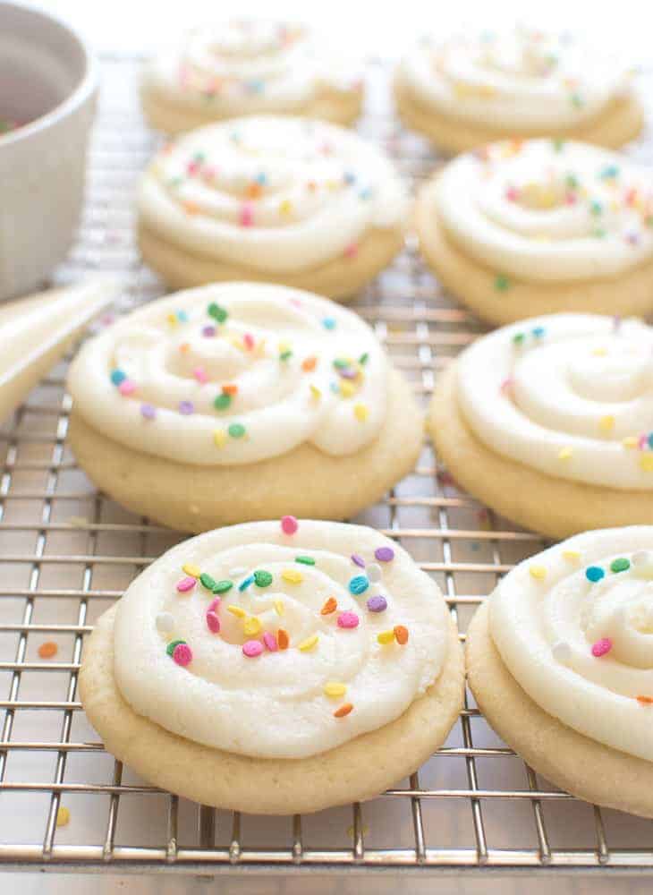 Greek Yogurt Sugar Cookies with pale cookies on a wire rack with white frosting and colored sprinkles on top.