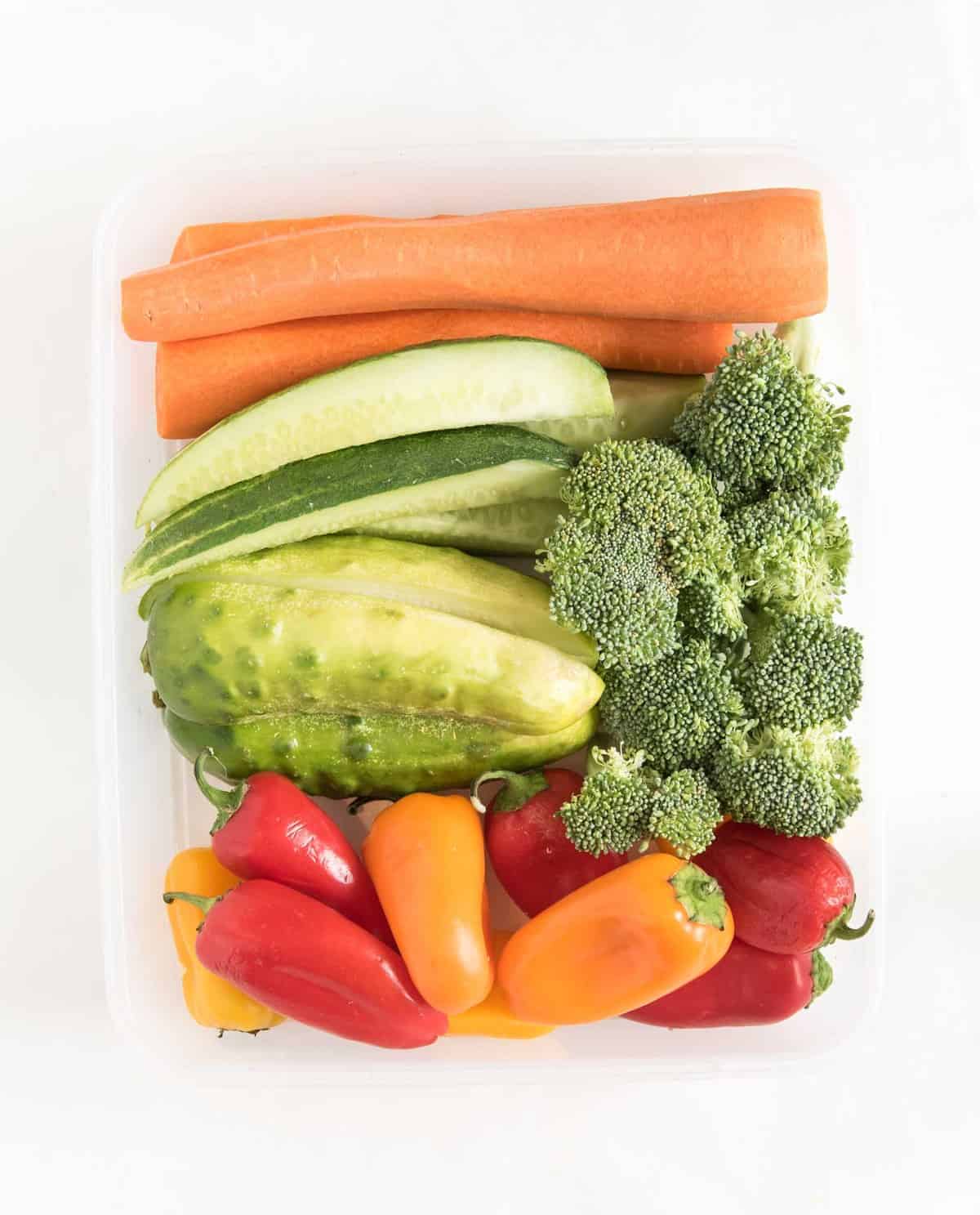 orange carrots, green broccoli and cucumbers, and multicolored peppers cut up in a clear plastic bowl.