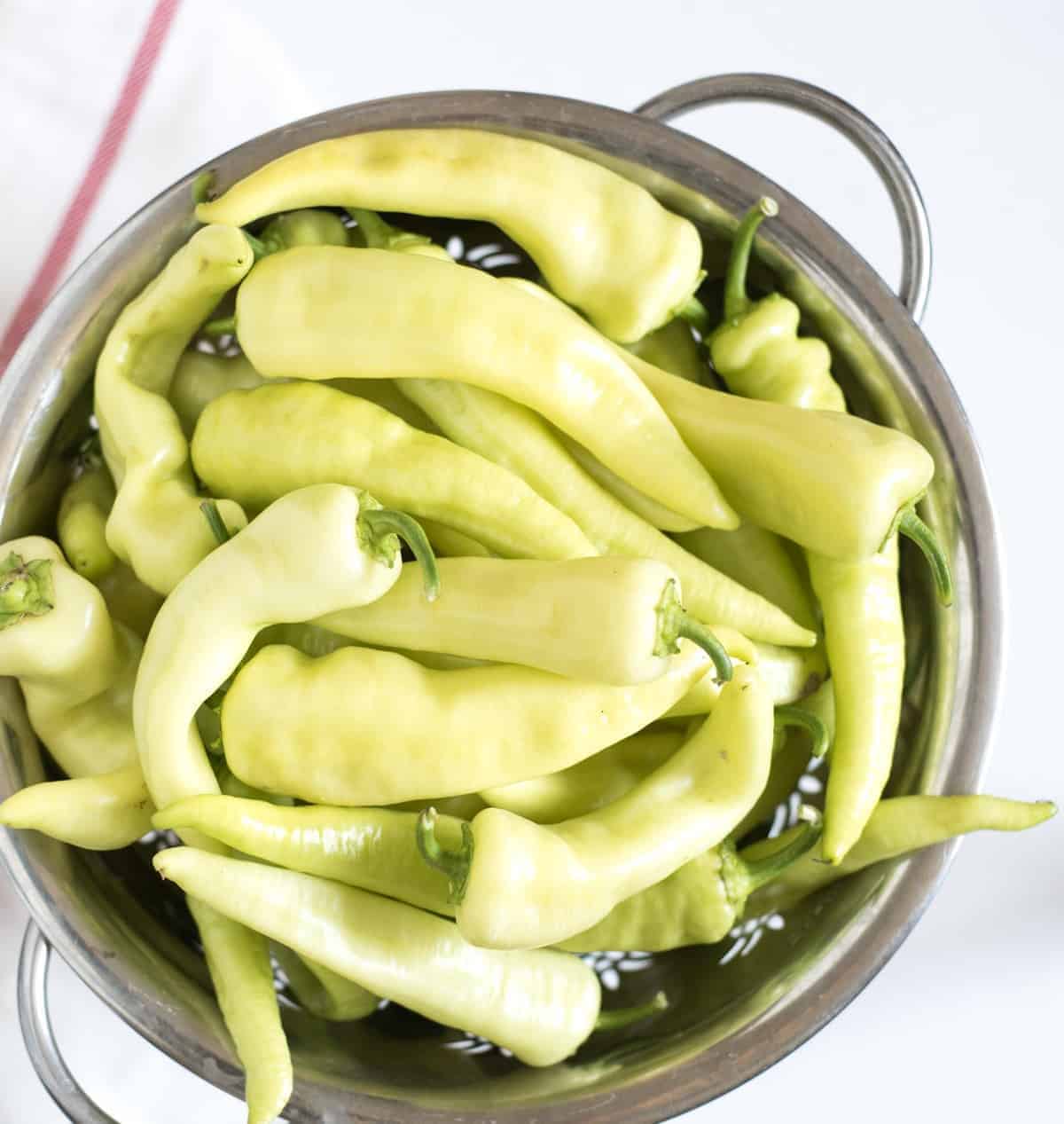 A metal colander filled with fresh yellow banana peppers sits on a light surface, perfect for those looking to learn how to pickle peppers.