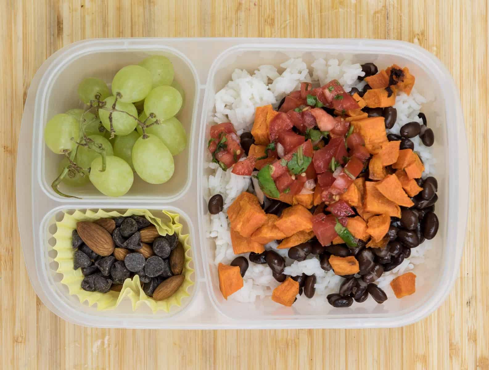 Top view of a lunch box with Rice + black beans + roasted sweet potatoes + fresh salsa, Almonds + chocolate chips, Grapes