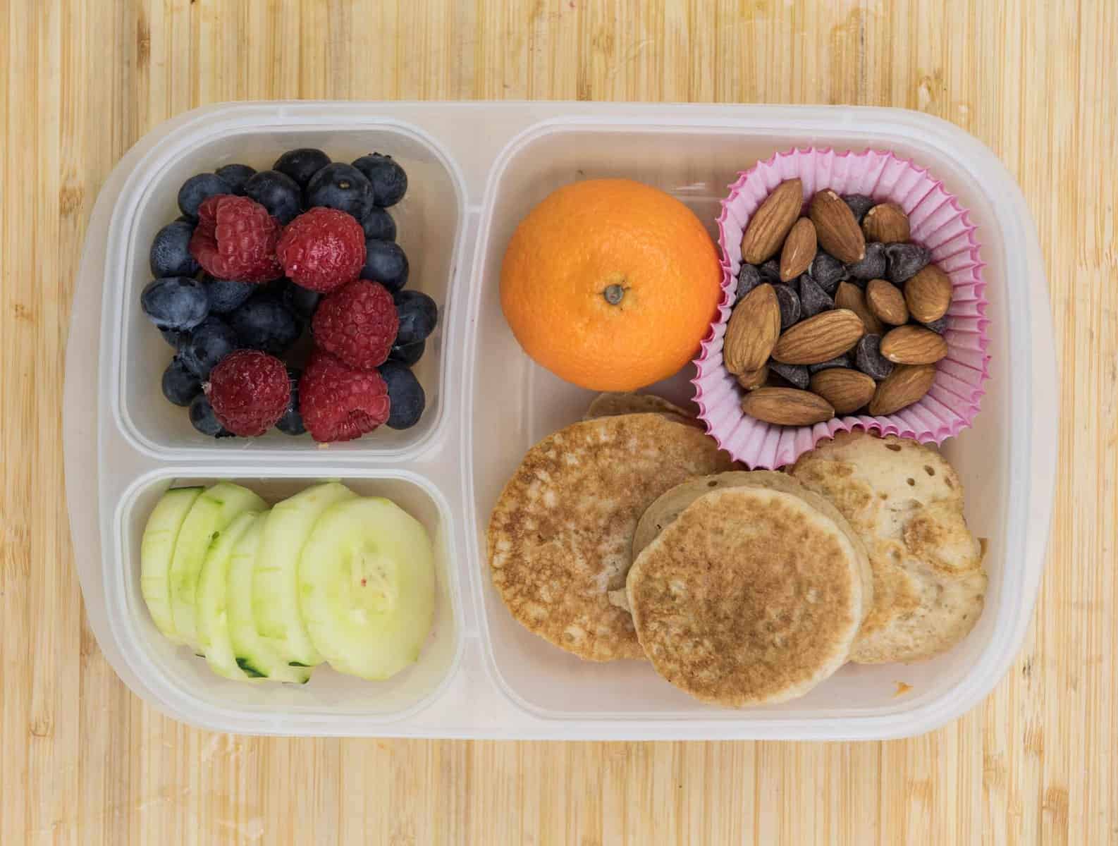 Top view of lunch box with mini pancakes, nuts and chocolate chips, fruit, and cucumber slices.