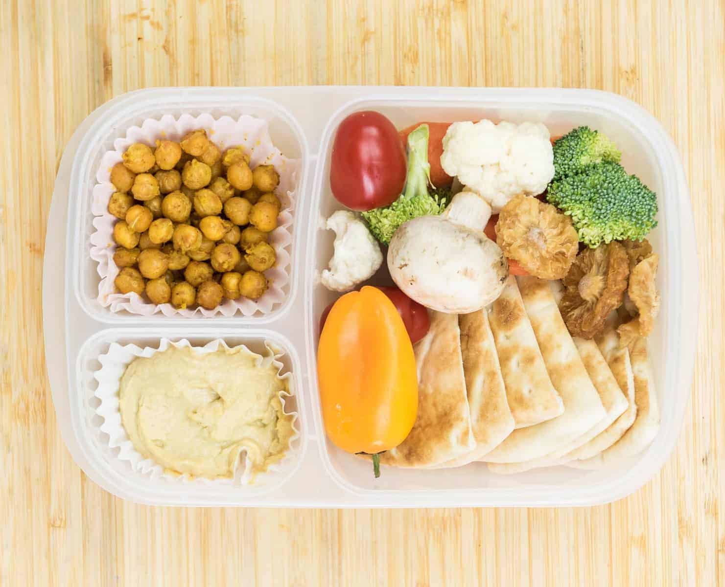 Top view of a lunch box with Hummus, Curry roasted chickpeas, Pita, Veggies such as peppers, tomatoes, carrots, mushrooms, broccoli, and/or cauliflower, and Dried pineapple.