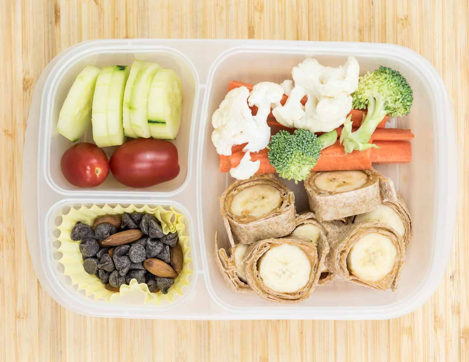 Top view of a lunch box with Banana Sushi, Carrots + tomatoes + cucumber + cauliflower and/or broccoli, and Chocolate chips + almonds.