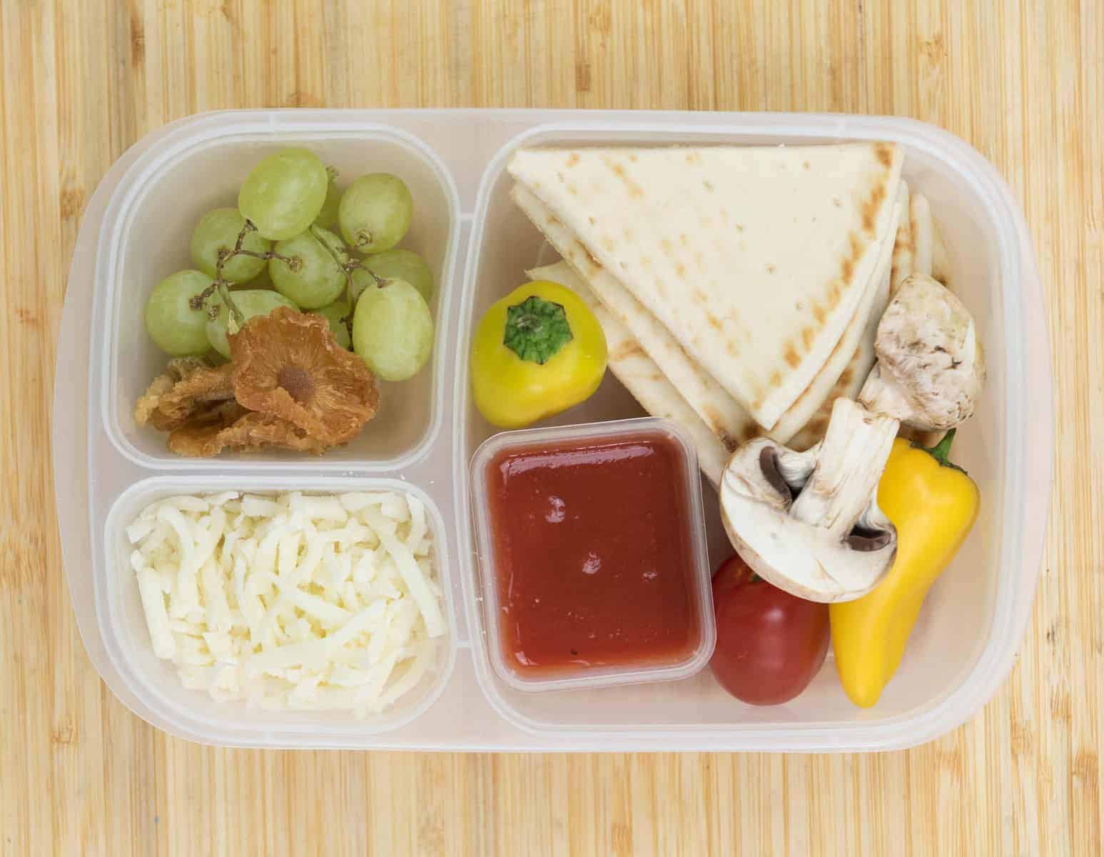 Top view of a lunch box with Pita, Tomatoes + mushrooms + bell peppers, Pizza sauce, Mozzarella cheese, and Dried pineapple + grapes