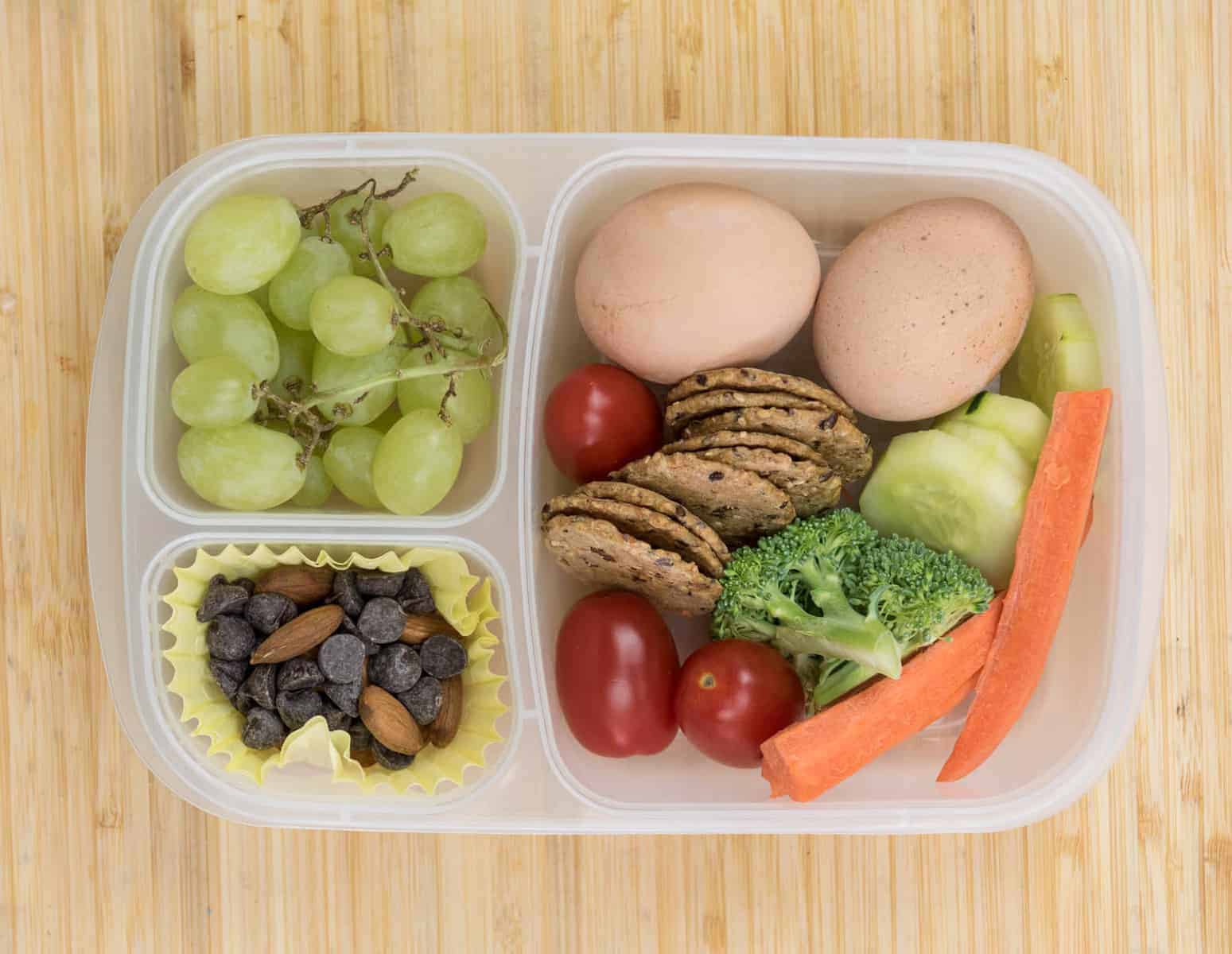 Top view of a lunch box with two boiled eggs, veggies, grapes, crackers, and nuts and chocolate chips.