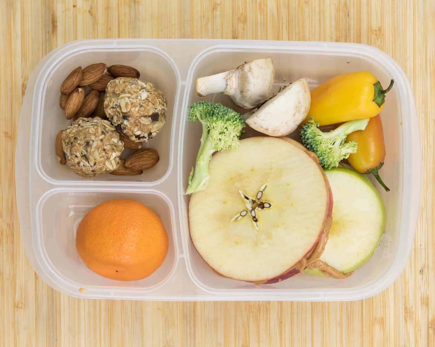 Top view of a lunch box with Apple sandwiches, Oranges, No-bake oatmeal bites + almonds, Veggies like peppers, broccoli, or mushrooms