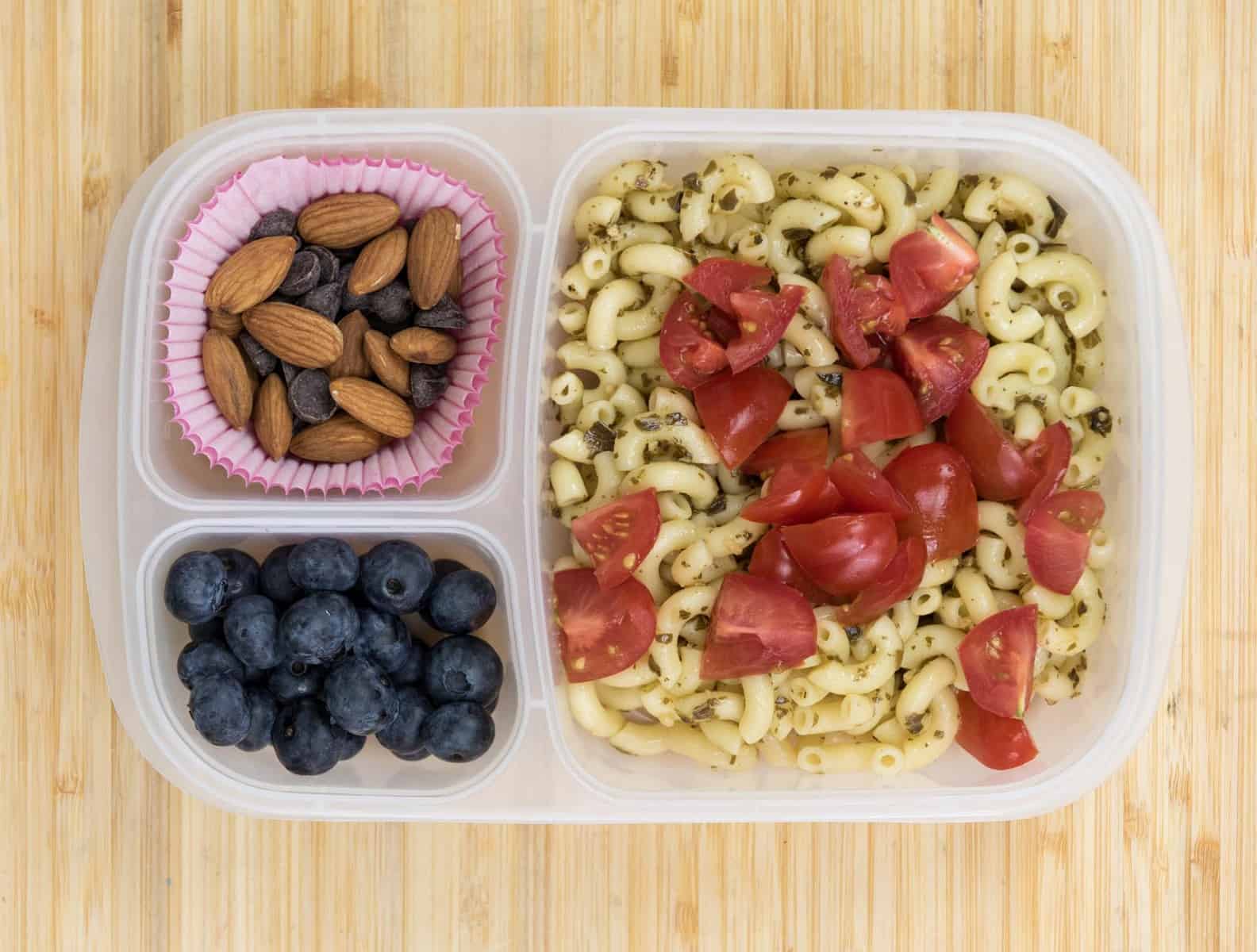 Top view of a lunch box with Cooked pasta + pesto + chopped tomato, Chocolate chips + almonds, and Blueberries